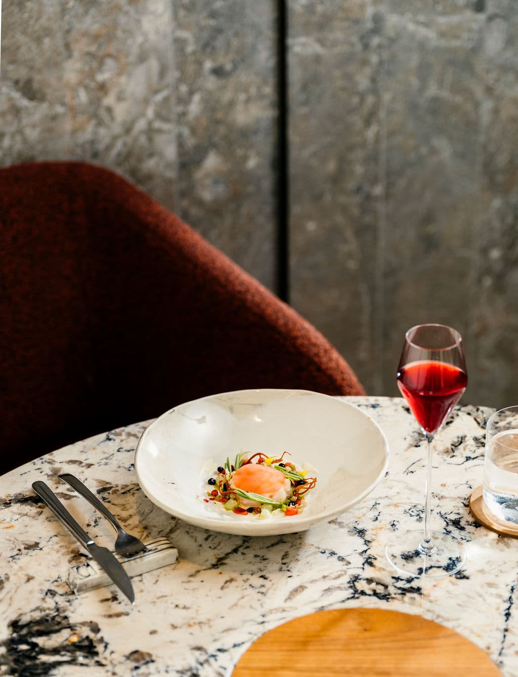 A white bowl of a refined dish served on a marble table with a glass of red wine.