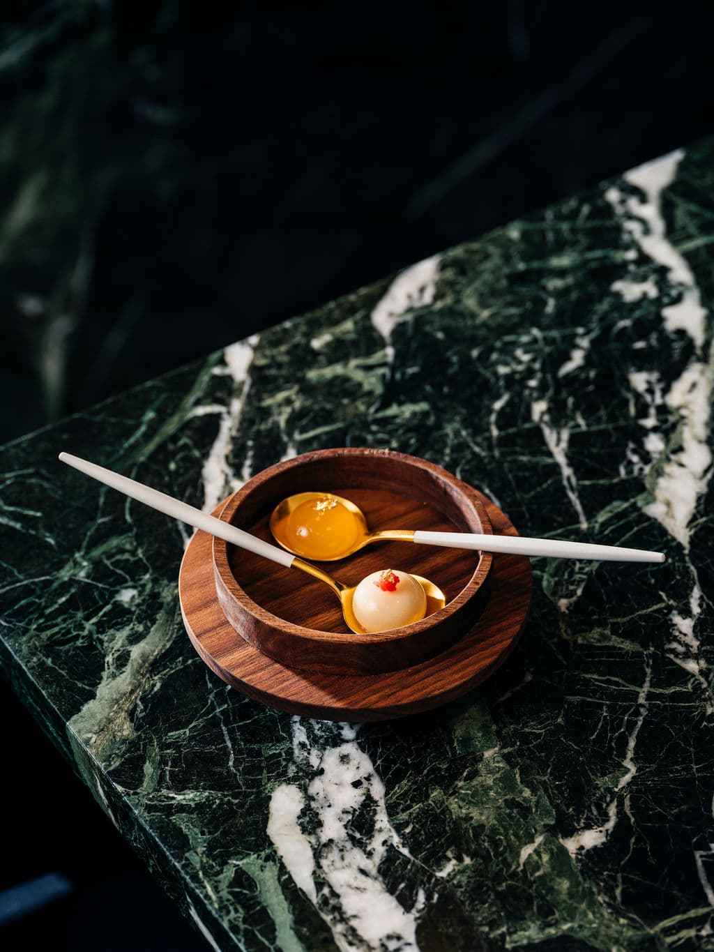 A wooden bowl with two golden spoons holding delicate bite-sized creations on a green marble table.