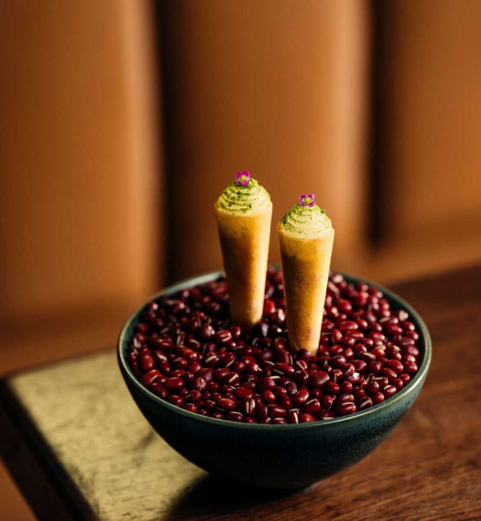 Two miniature savory cones topped with green herbs and tiny edible flowers, presented in a bowl of red beans.