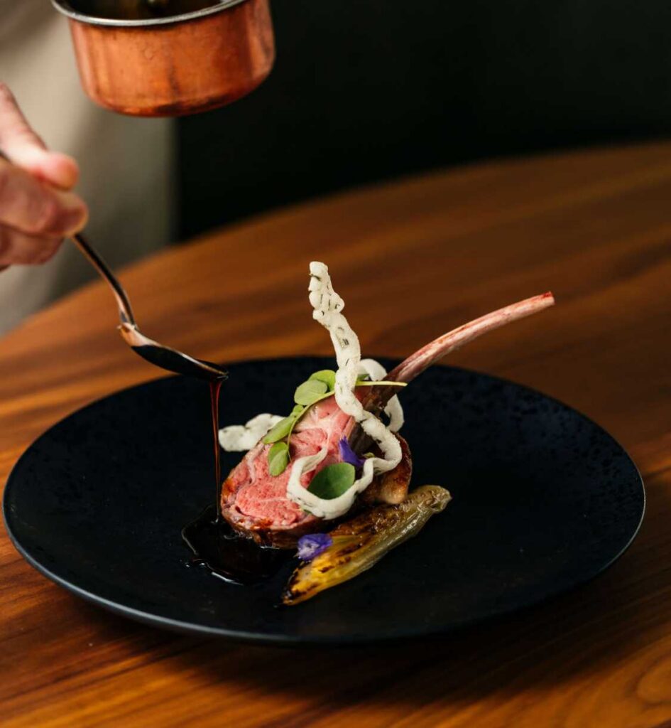Lamb chop plated with greens and sauce being poured from a spoon.