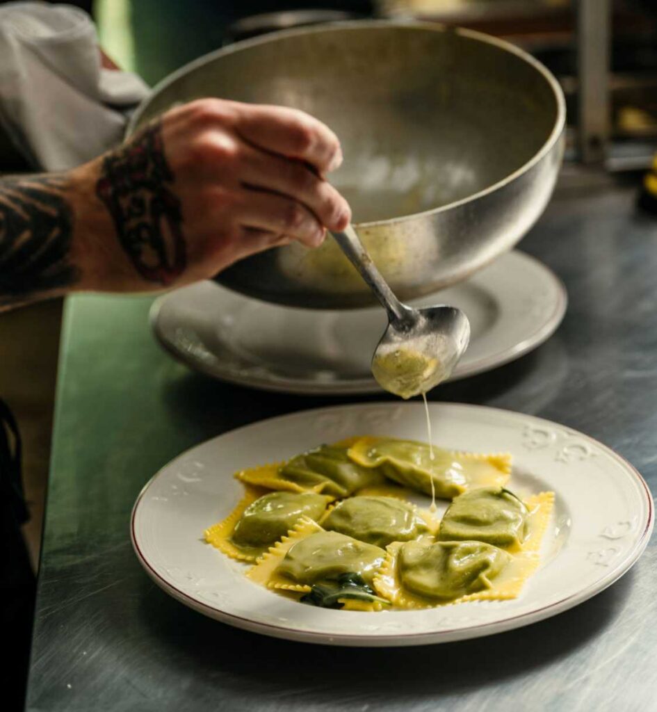 Chef ladling sauce over green ravioli on a white plate.