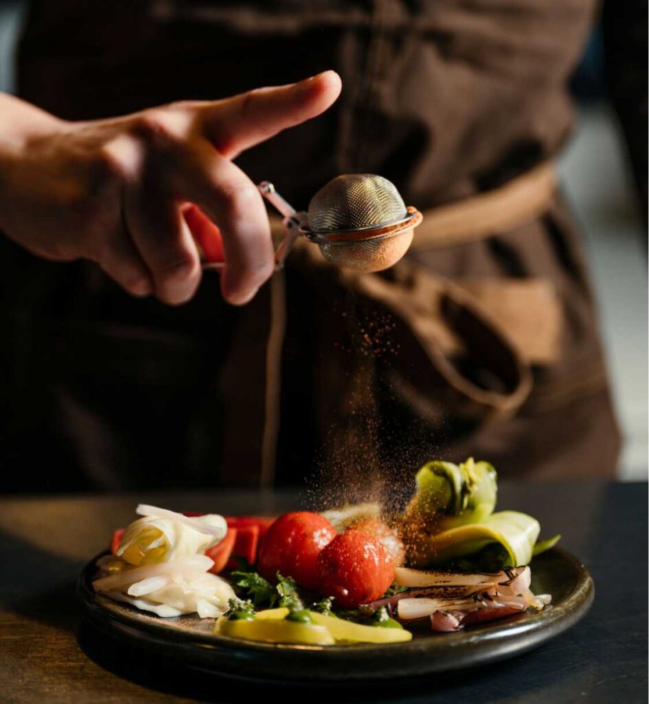 Chef dusting seasoning over a plate of colorful vegetables, including tomatoes, greens, and pickled slices.