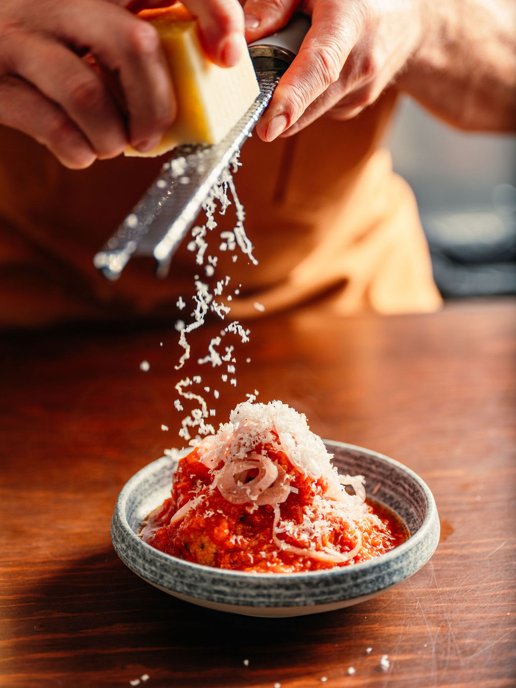 Du fromage tombe d'une râpe sur une boulette en sauce tomate garnie d'oignon émincé, dans un bol bordé de bleu.