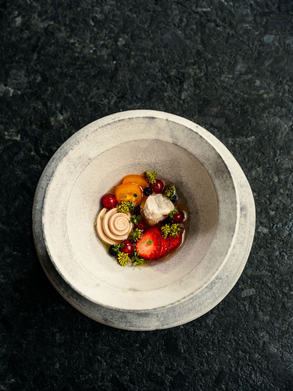Petits fruits, tomates tranchées et fleurs de fenouil en bouillon clair, dans un bol pâle sur pierre sombre.