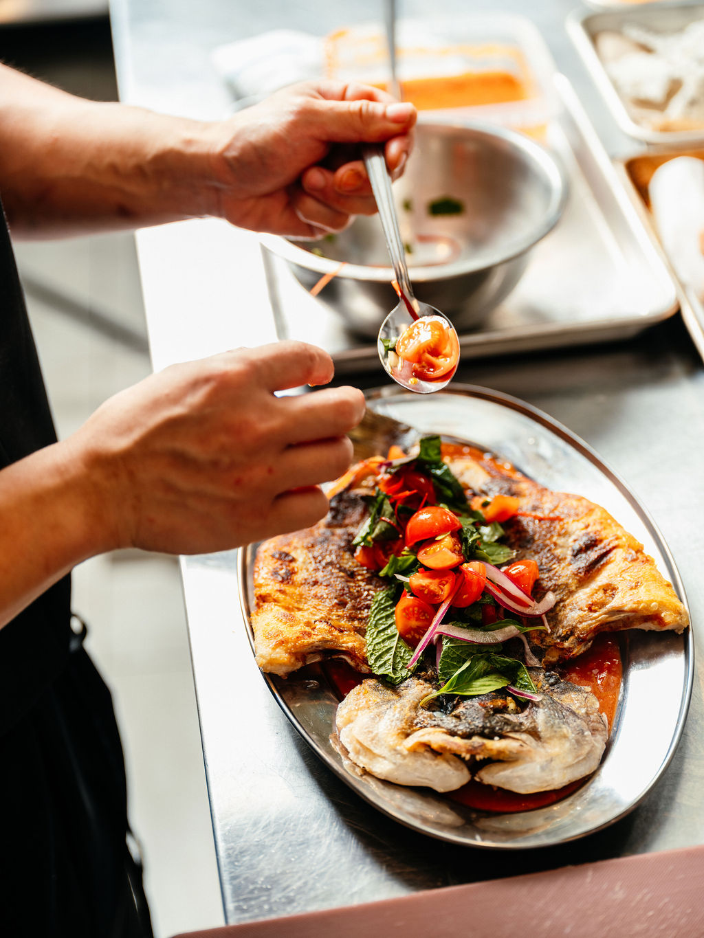 Des mains déposent des tomates cerises sur un poisson entier croustillant, menthe et basilic, plat en acier.