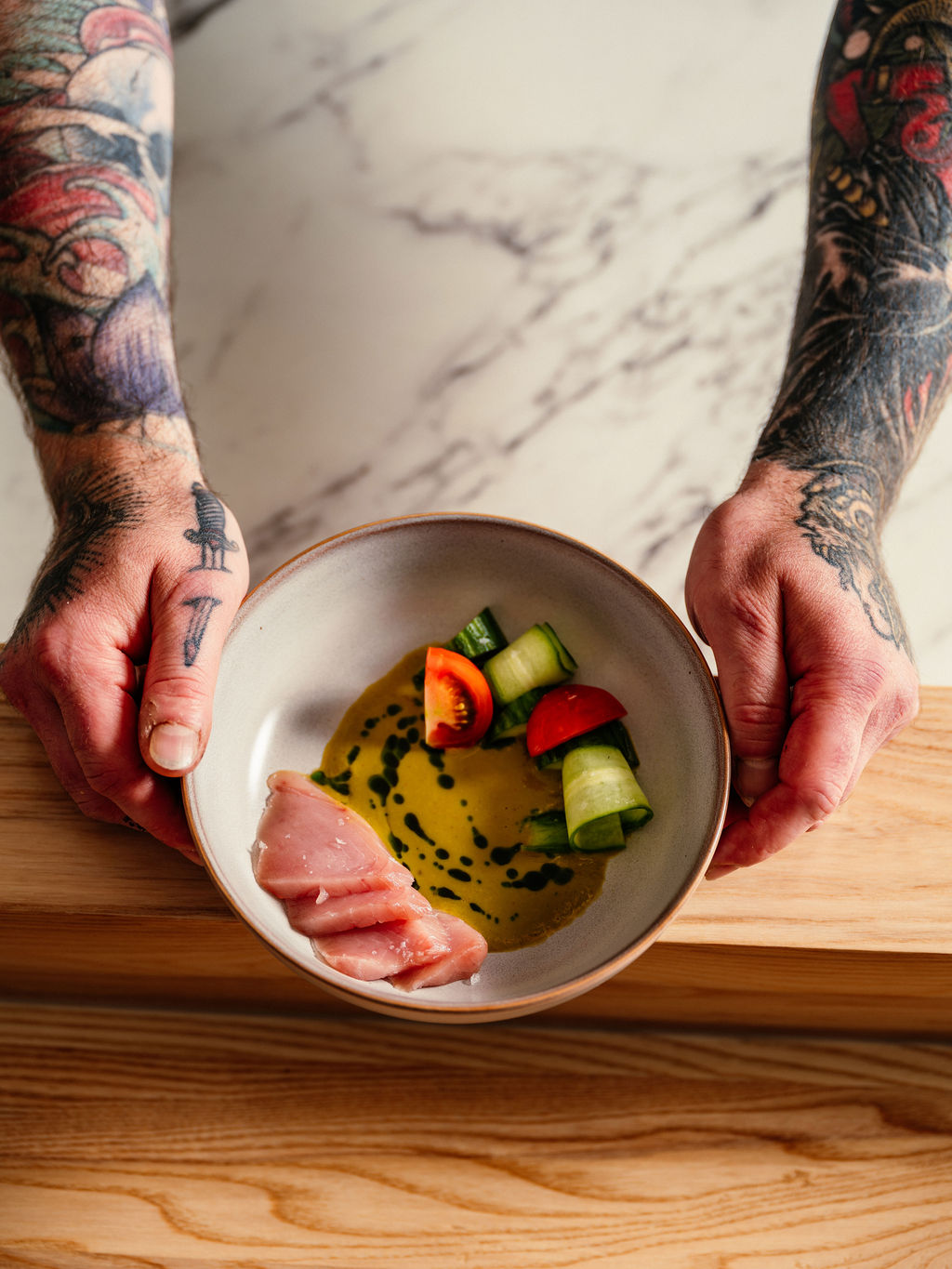 Des mains tatouées déposent un bol de poisson cru, rouleaux de concombre, tomate et sauce jaune-vert, sur bois.