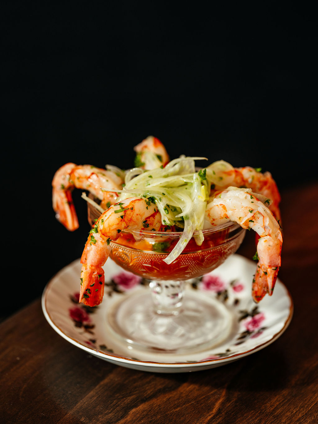 Cinq crevettes accrochées à une coupe de verre pressé garnie de sauce cocktail et de fenouil, soucoupe fleurie.