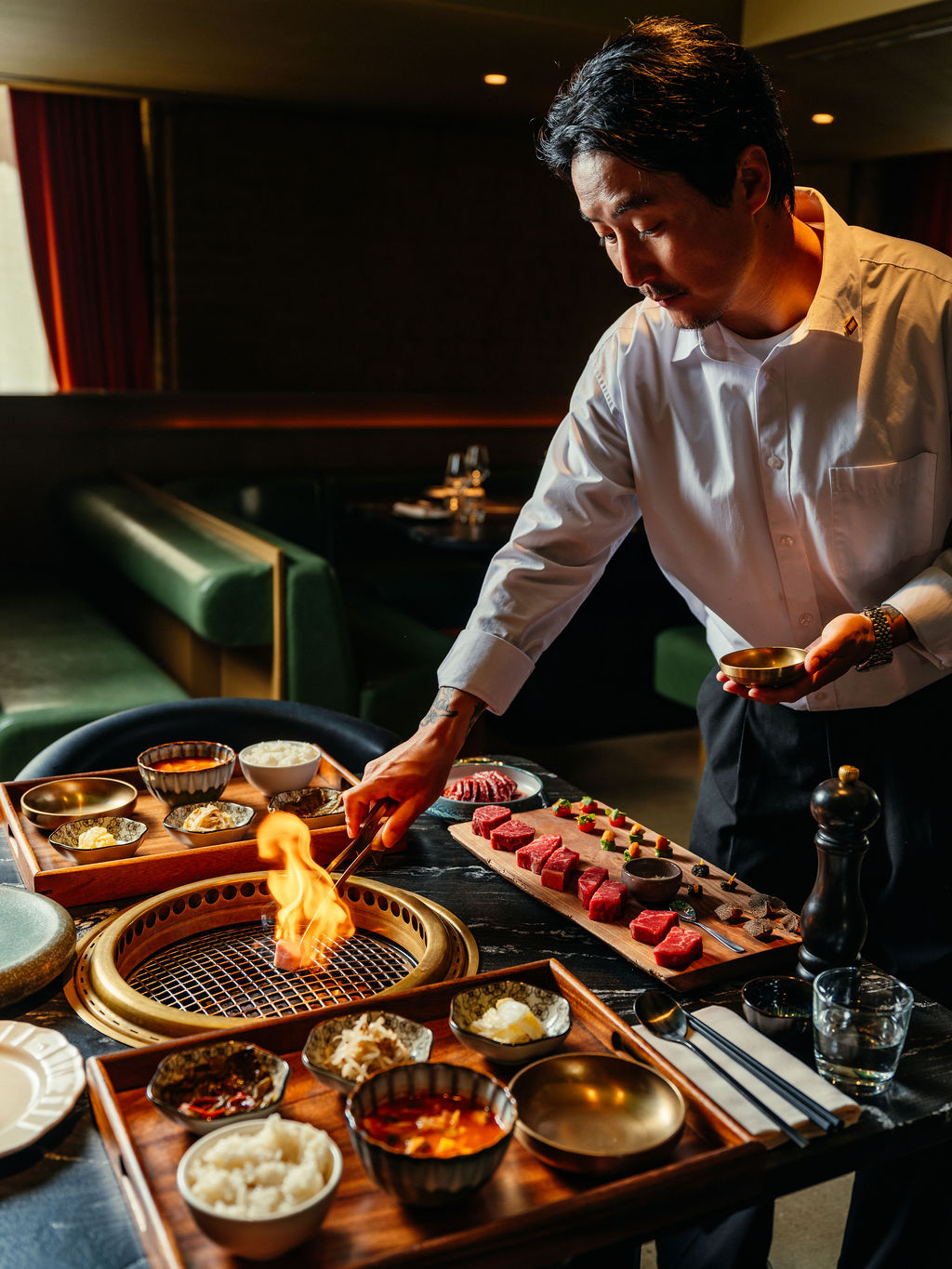 Un chef se penche sur un gril de table enflammé, entouré de bols de laiton, de banchan et de bœuf cru.