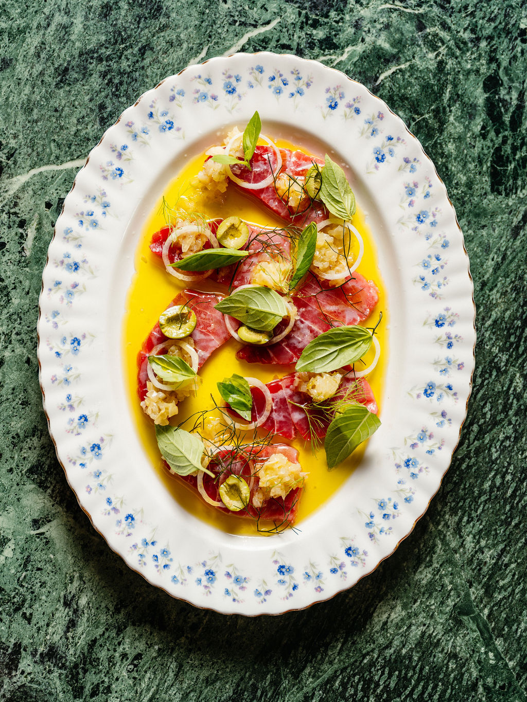 Fines tranches de poisson cru, oignon, olives vertes et basilic dans une huile dorée, plat ovale fleuri.