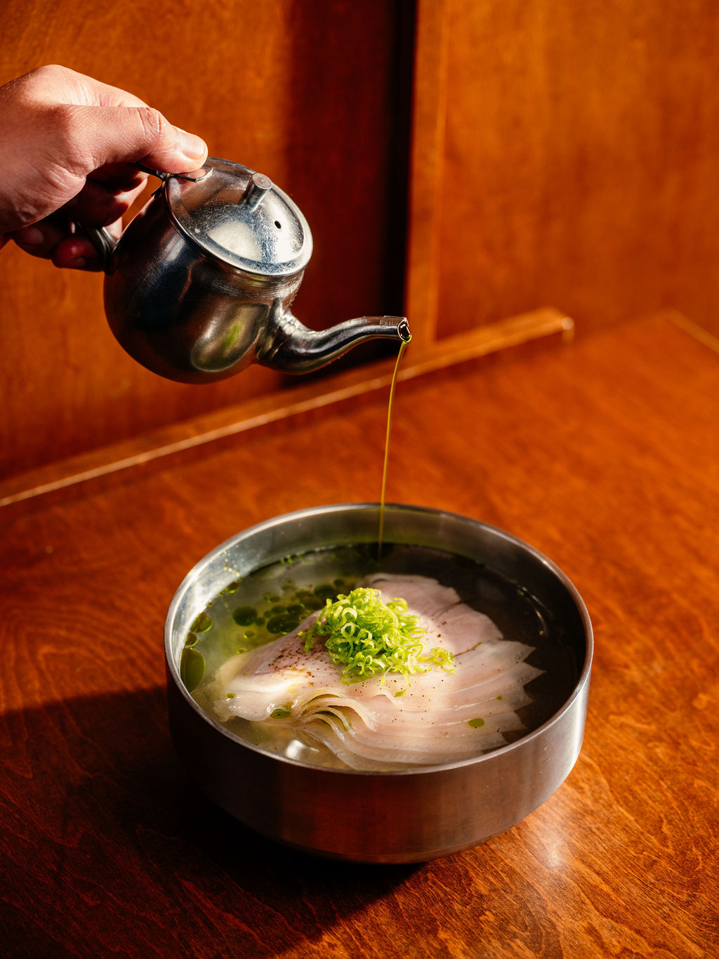De l'huile verte coule d'un pot de métal sur des tranches de poisson et de l'oignon vert, bol de laiton.