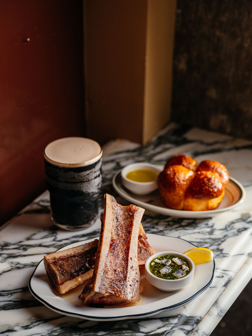 Os à moelle rôtis, anchois sur sauce verte et citron, près d'une stout et de brioches dorées, sur marbre.