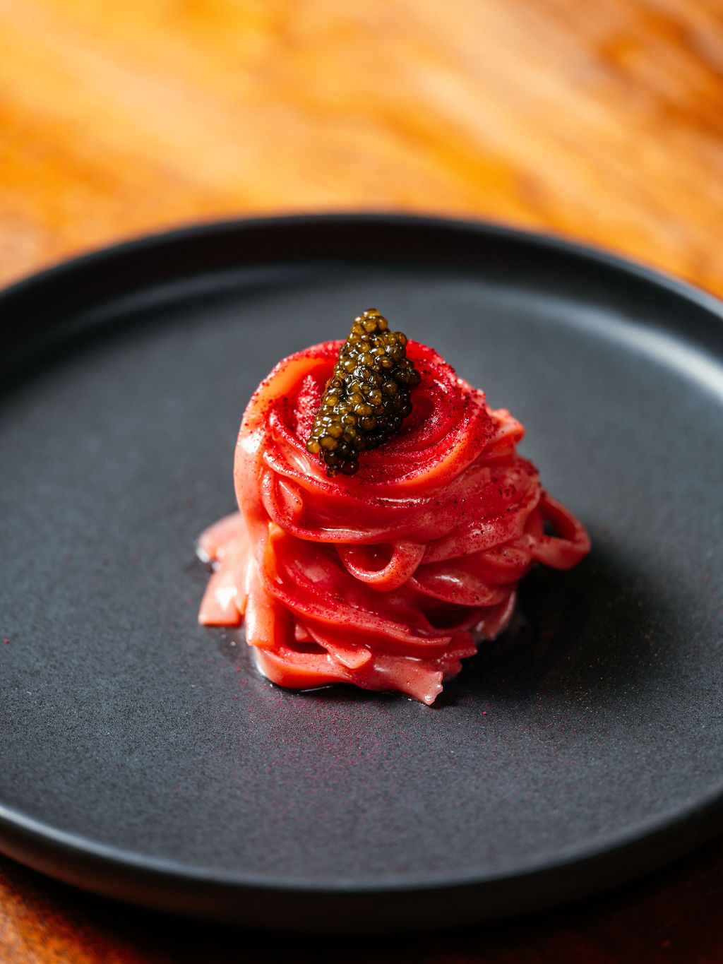Un nid de tagliolini rosés couronné d'une quenelle de caviar, dans une assiette noire mate sur bois clair.