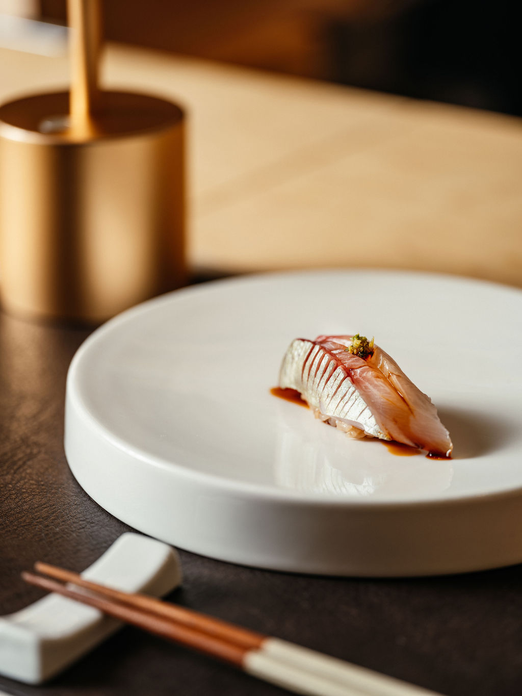 Une bouchée de nigiri au poisson à peau argentée et wasabi râpé, dans une assiette ronde, près de baguettes.